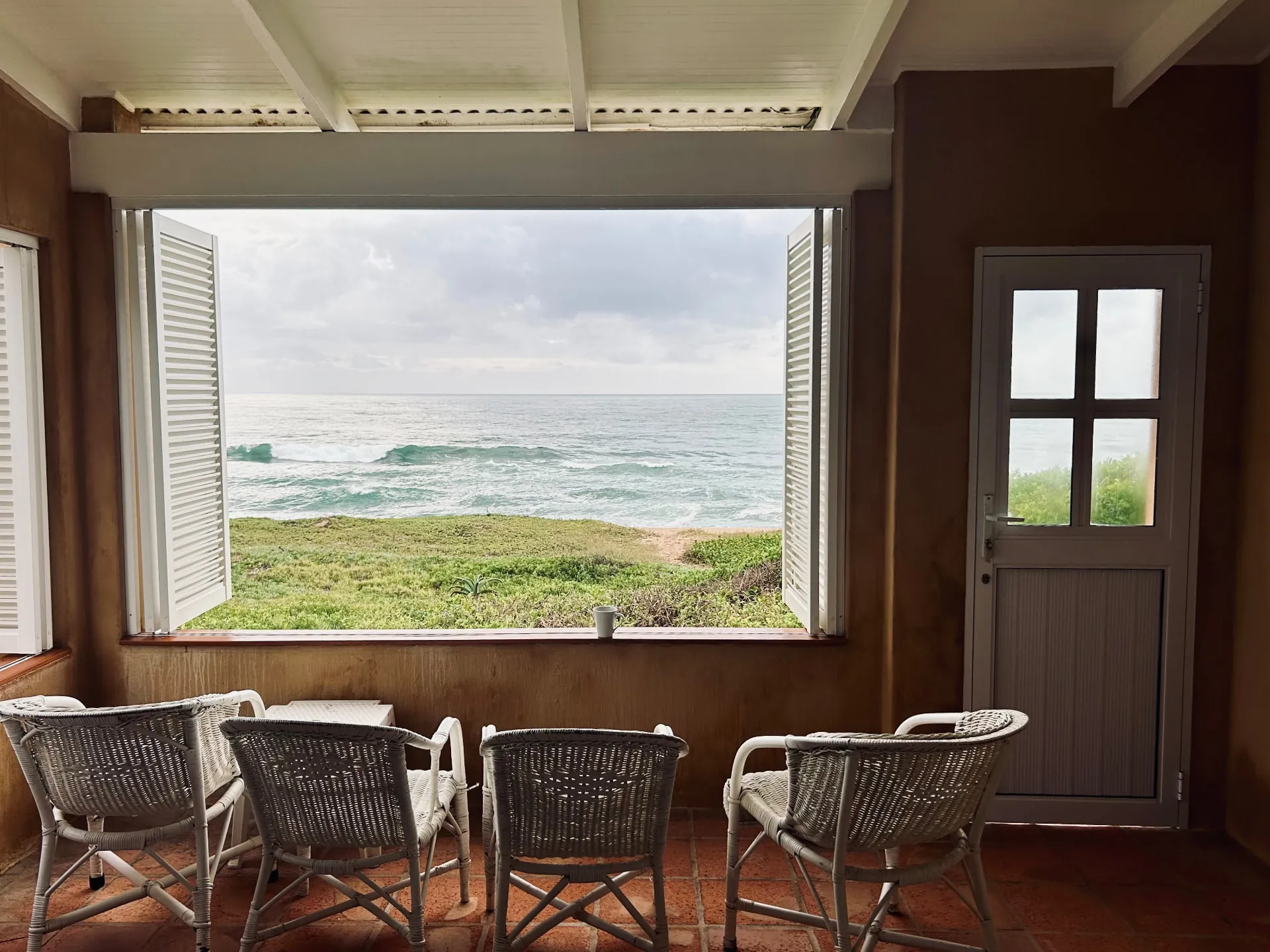Open shuttered window at The Beach Cottage looking over dune grass to breaking waves, with wicker chairs on a terracotta tiled veranda